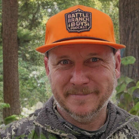 Man wearing an orange cap with 'Battle Branch Boys Podcast' logo in a forest setting